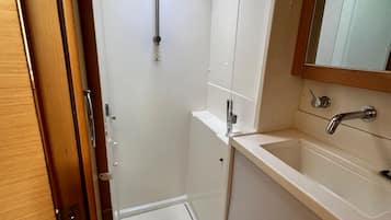 Un espacio de ducha pequeño y cerrado, con una ventana, un lavamanos con espejo y una puerta.