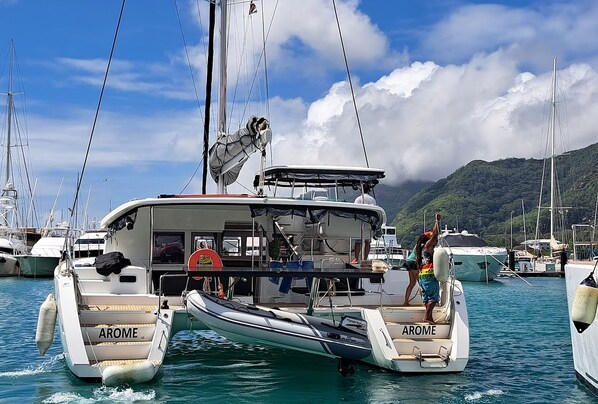 A white boat with 'AROME' written on the side, docked at a marina with other boats and a mountainous backdrop.