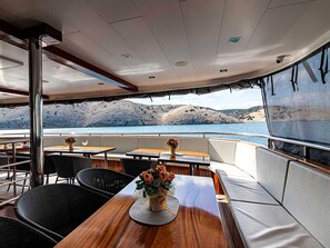 A boat's dining area with tables, chairs, and a view of the sea and hills.