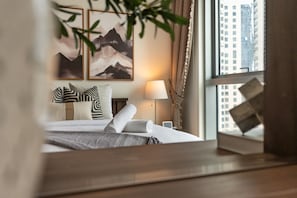 Une chambre d’hôtel avec un lit, des oreillers, une lampe de chevet, une fenêtre avec des rideaux et des œuvres d’art encadrées accrochées au mur.