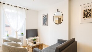 A living room with a grey sofa, two armchairs, a coffee table with a plant, a TV on a white stand, and a mirror on the wall.