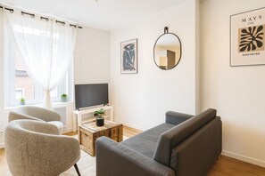 A living room with a grey sofa, two armchairs, a coffee table with a plant, a TV on a white stand, and a mirror on the wall.