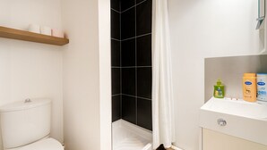 A bathroom with a toilet, a shower with black tiles, a white curtain, and a shelf with toiletries.
