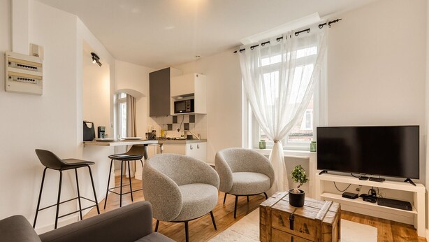 A modern living room with a sofa, two armchairs, a coffee table, and a TV. There is a kitchen in the background with a microwave and cabinets.
