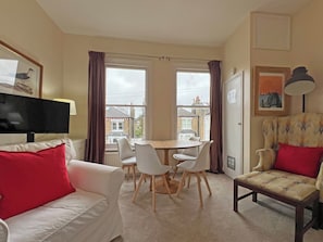 Ein Wohnzimmer mit einem weißen Sofa, einem gemusterten Sessel, einem Esstisch mit weißen Stühlen und einem Fernseher.