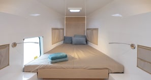 A narrow, elongated room with a bed at the center, a window on one side, and a shower area on the other.