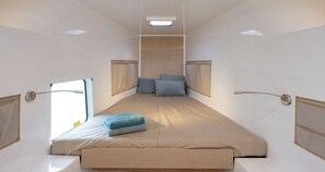 A narrow, elongated room with a bed at the center, a window on one side, and a shower area on the other.