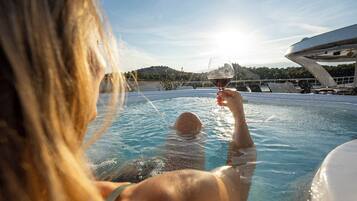 Une personne dans une piscine, tenant un verre de vin, avec un ciel dégagé et un bâtiment en arrière-plan.