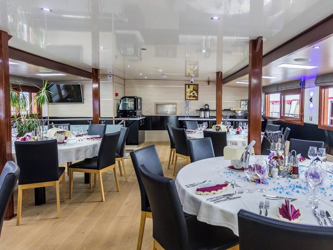 Un restaurant avec des tables rondes, des chaises noires et un espace cuisine à l’arrière-plan.