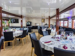 Ein Restaurant mit runden Tischen, schwarzen Stühlen und einer Küche im Hintergrund.