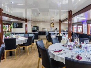 A restaurant with round tables, black chairs, and a kitchen area in the background.
