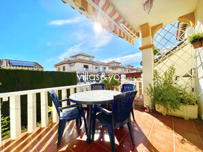 En terrasse med et bord og stole, et hvidt gelænder og udsigt til bygninger i baggrunden.