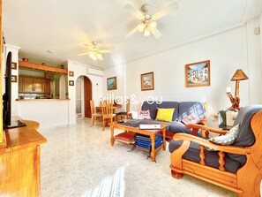 Ein großzügiges Wohnzimmer mit einer Couch, Sesseln, einem Couchtisch und einem Essbereich. Es gibt Deckenventilatoren, eine durch einen Bogen sichtbare Küche und gerahmte Bilder an den Wänden.