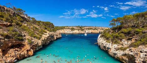 A coastal beach with clear turquoise water, rocky cliffs, and a boat in the distance.