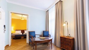 Habitación de hotel con suelo de madera, una mesa redonda con sillas, mesita de noche con lámpara y un armario.