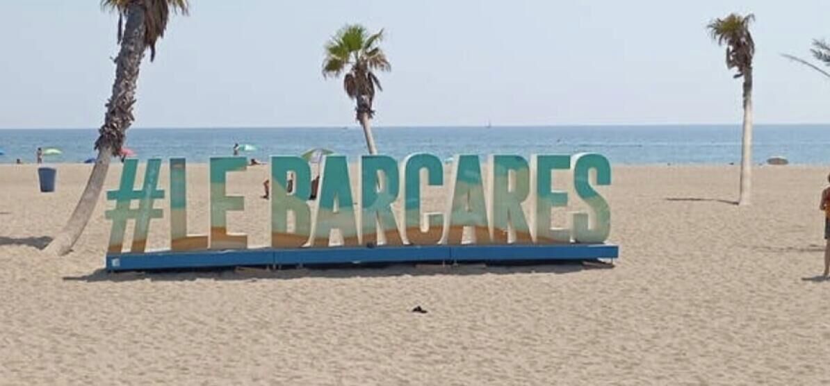 Een strand met een groot bord waarop staat '#LEBARCARES' en palmbomen op de achtergrond.