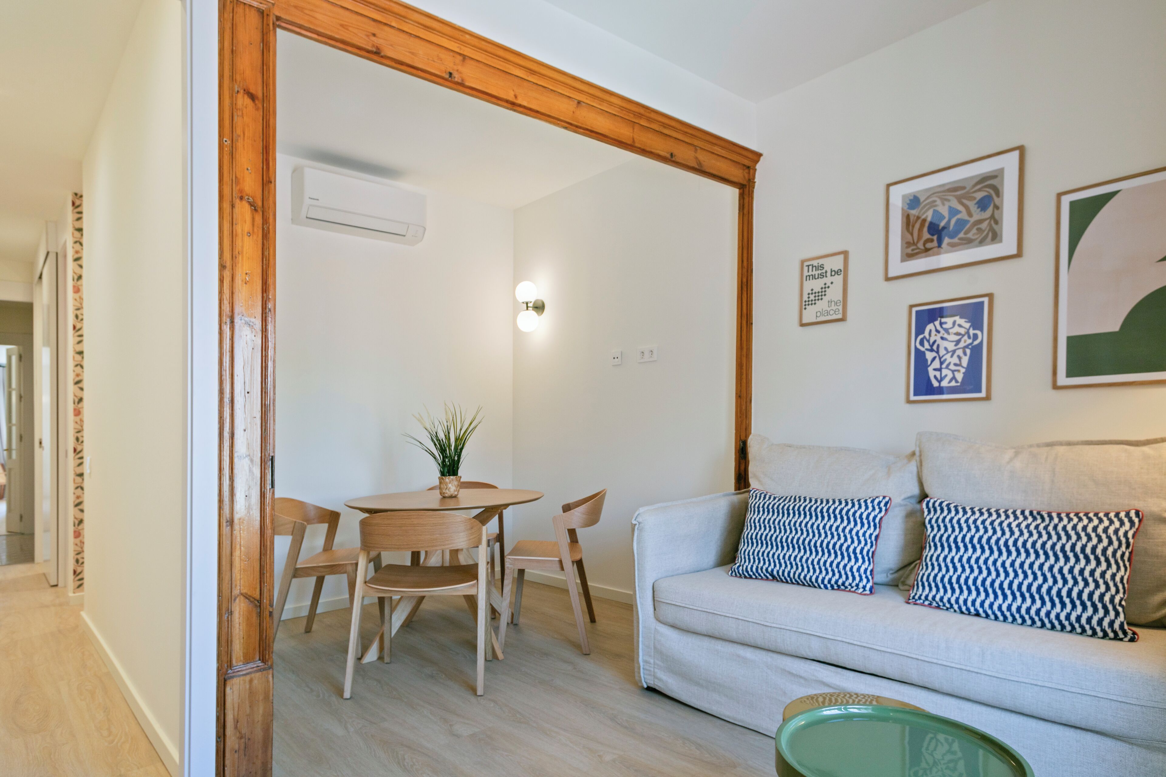 Ein Zimmer mit einer holzrahmenverzierten Tür, einem Esstisch mit Stühlen, einer Couch mit gemusterten Kissen und an der Wand befestigter Kunst.
