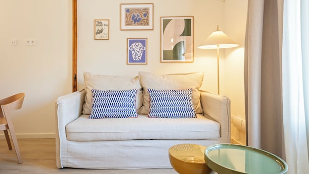 A living room with a sofa, two patterned pillows, a lamp, a chair, and three framed artworks on the wall.
