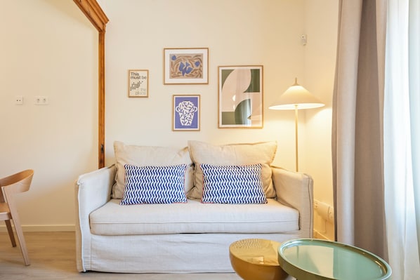 A living room with a sofa, two patterned pillows, a lamp, a chair, and three framed artworks on the wall.