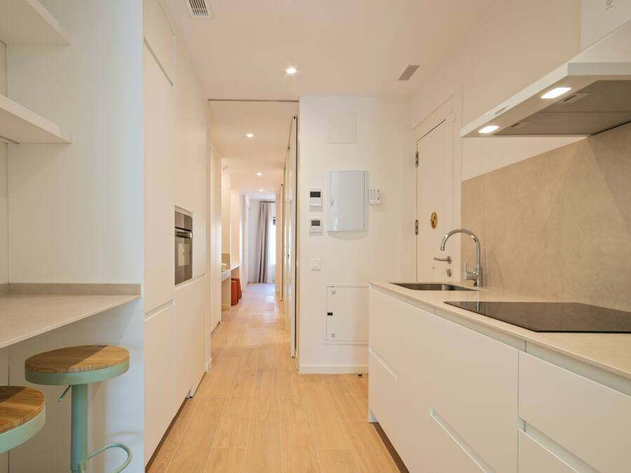 A narrow kitchen with white cabinetry, a stainless steel sink, and a wooden dining table.