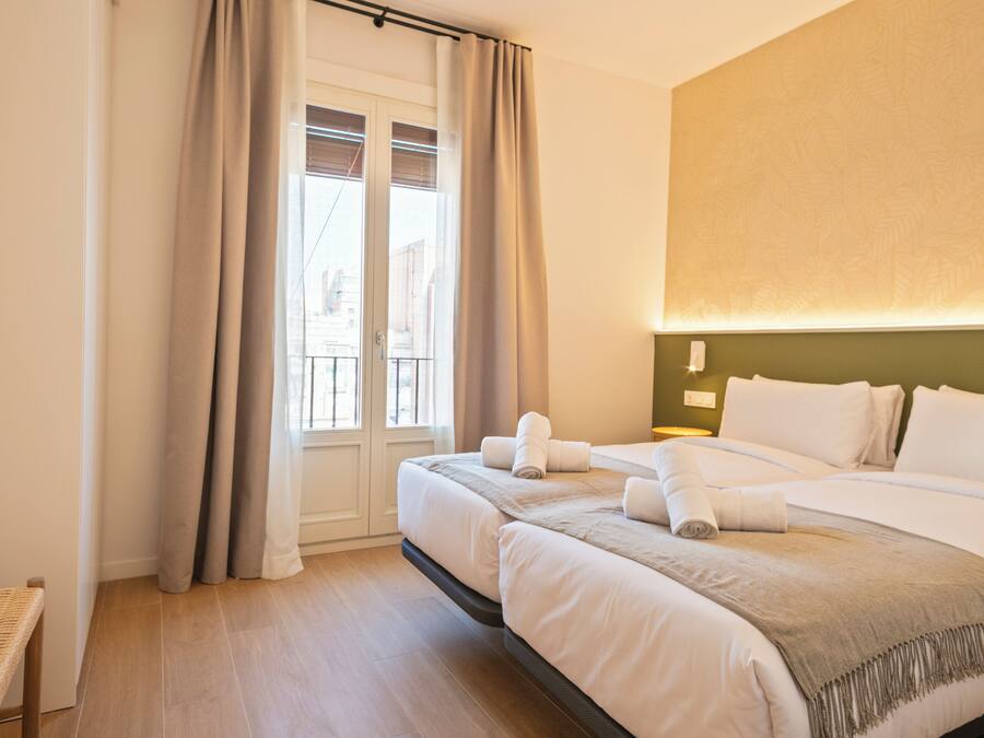A hotel room with two beds, a large window with curtains, a wall-mounted light, and a wooden floor.