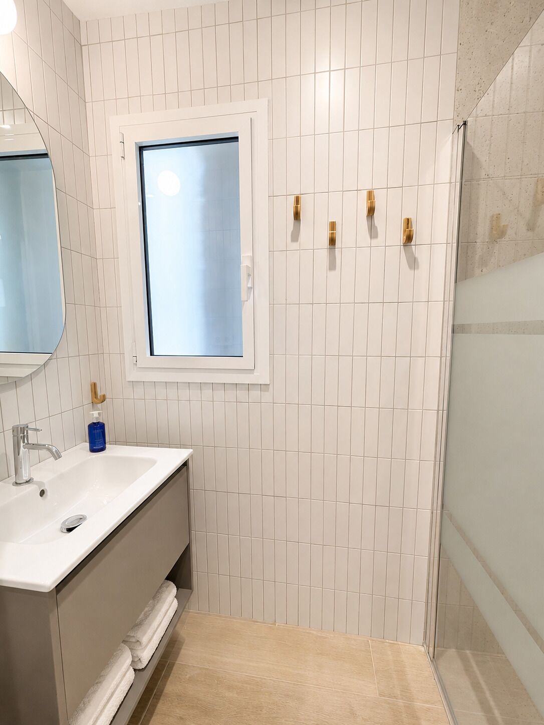 A bathroom with a white tiled wall, a window, a sink with a mirror, and a shower area.