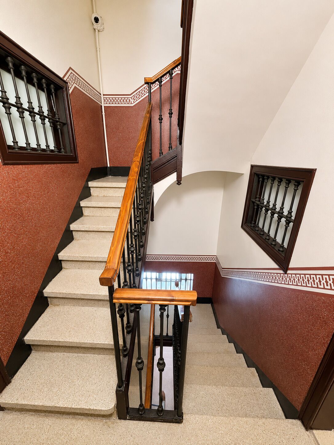 Eine Treppe mit hölzernem Geländer und schwarzen Spindeln, die zu einer Zwischenebene mit einem kleinen Fenster führt.