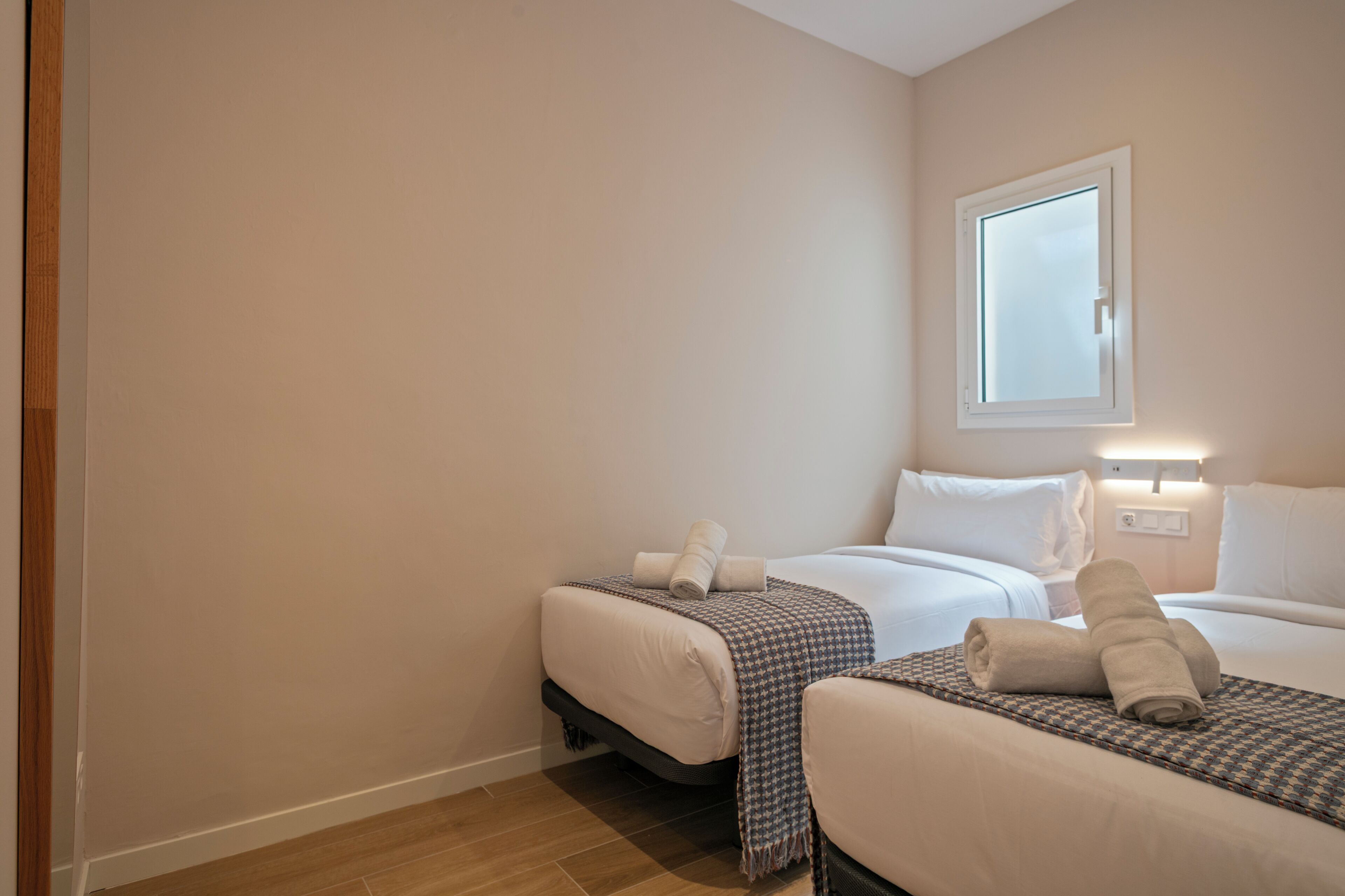 A hotel room with two beds, a window, and a wall-mounted light.