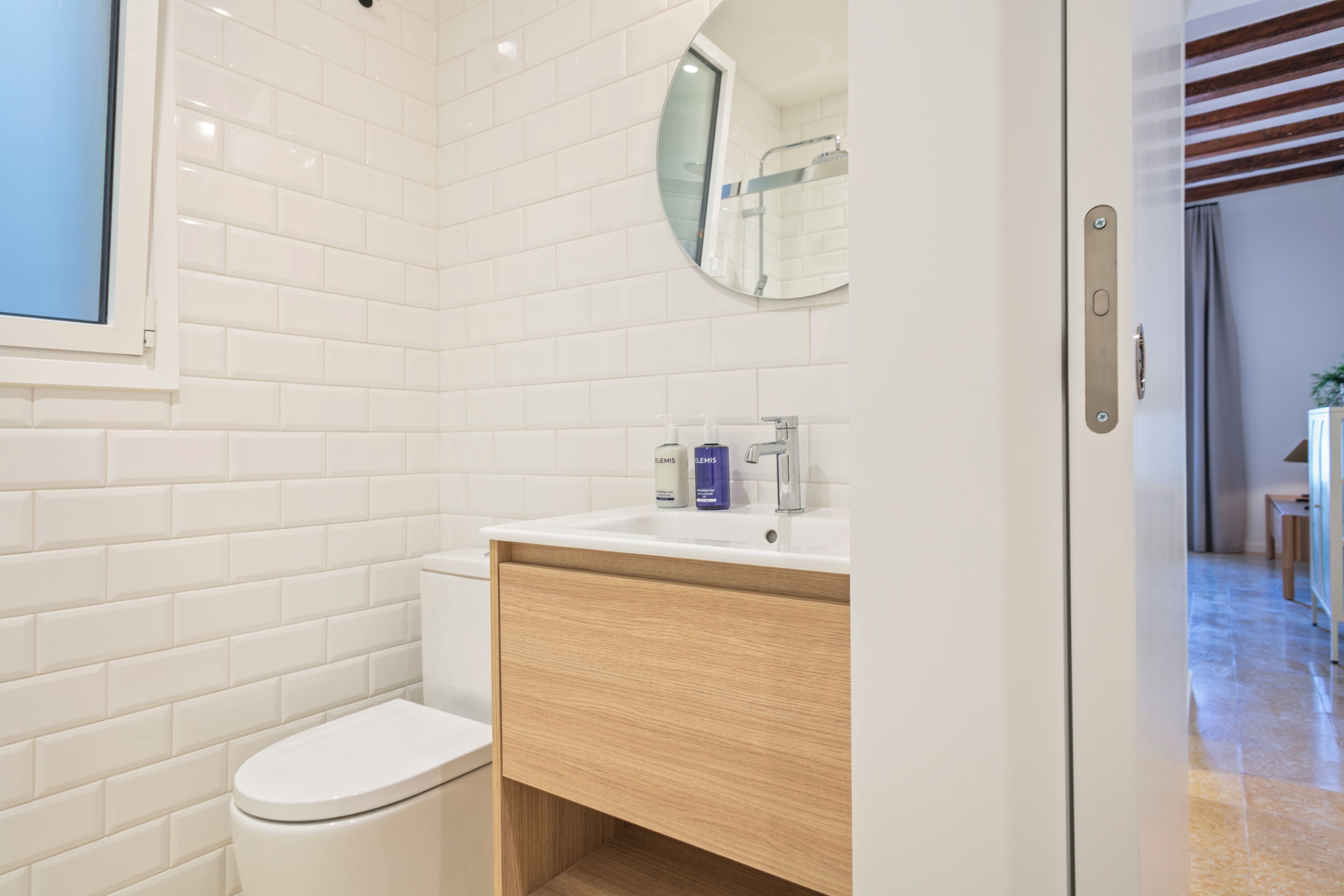 Une salle de bain moderne avec des carreaux de métro blancs, des toilettes blanches, un meuble-lavabo en bois et un miroir au-dessus du meuble-lavabo.