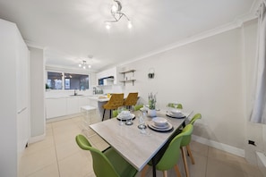 Un comedor moderno con una mesa de madera dispuesta para cuatro personas, rodeado de sillas verdes. Se ve una cocina con armarios blancos y una planta colgante al fondo.
