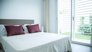 Un dormitorio con una cama grande, dos almohadas, una mesita de noche y un ventanal corredero que da a un balcón con follaje.