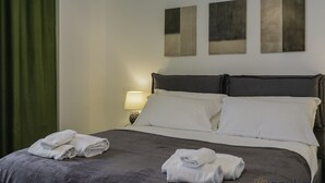 En snyggt bäddad säng med vita lakan och prydligt vikta handdukar, en sänglampa och tre abstrakta tavlor på väggen.