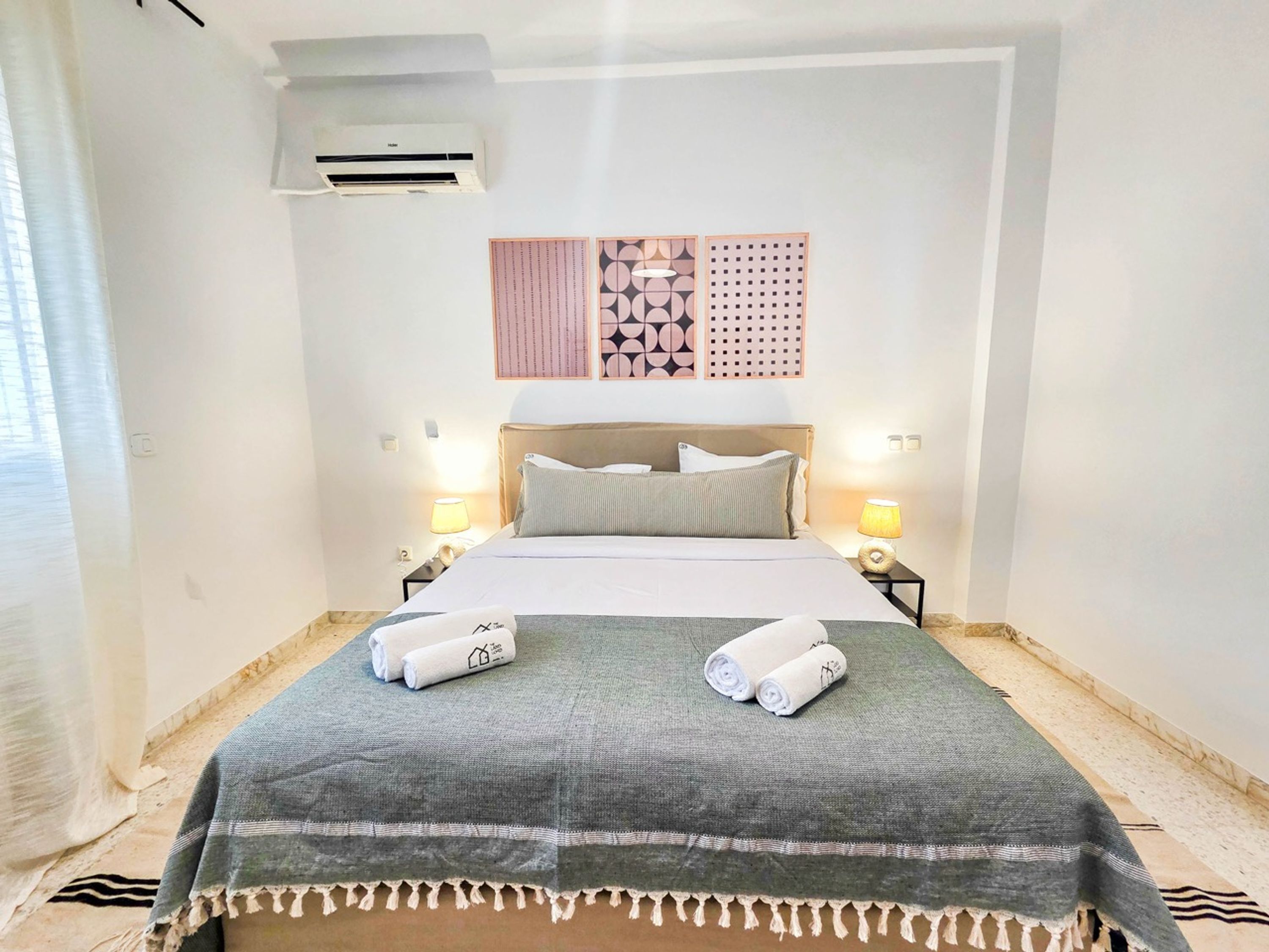 A hotel room with a bed, two bedside lamps, a wall-mounted air conditioner, and a decorative wall panel.