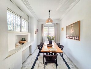 Un espace repas doté d’un sol en marbre, d’une table en bois, de chaises en osier et d’un luminaire suspendu.