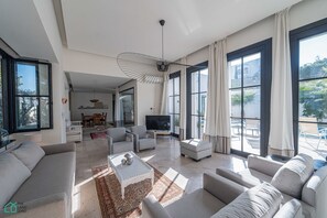 Ein geräumiges Wohnzimmer mit großem Fernseher, bequemen Sitzmöglichkeiten und großen Fenstern.
