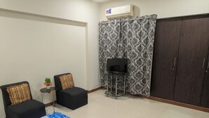 A small living area with a TV on a stand, two chairs with cushions, a small table with a plant, and a patterned curtain.