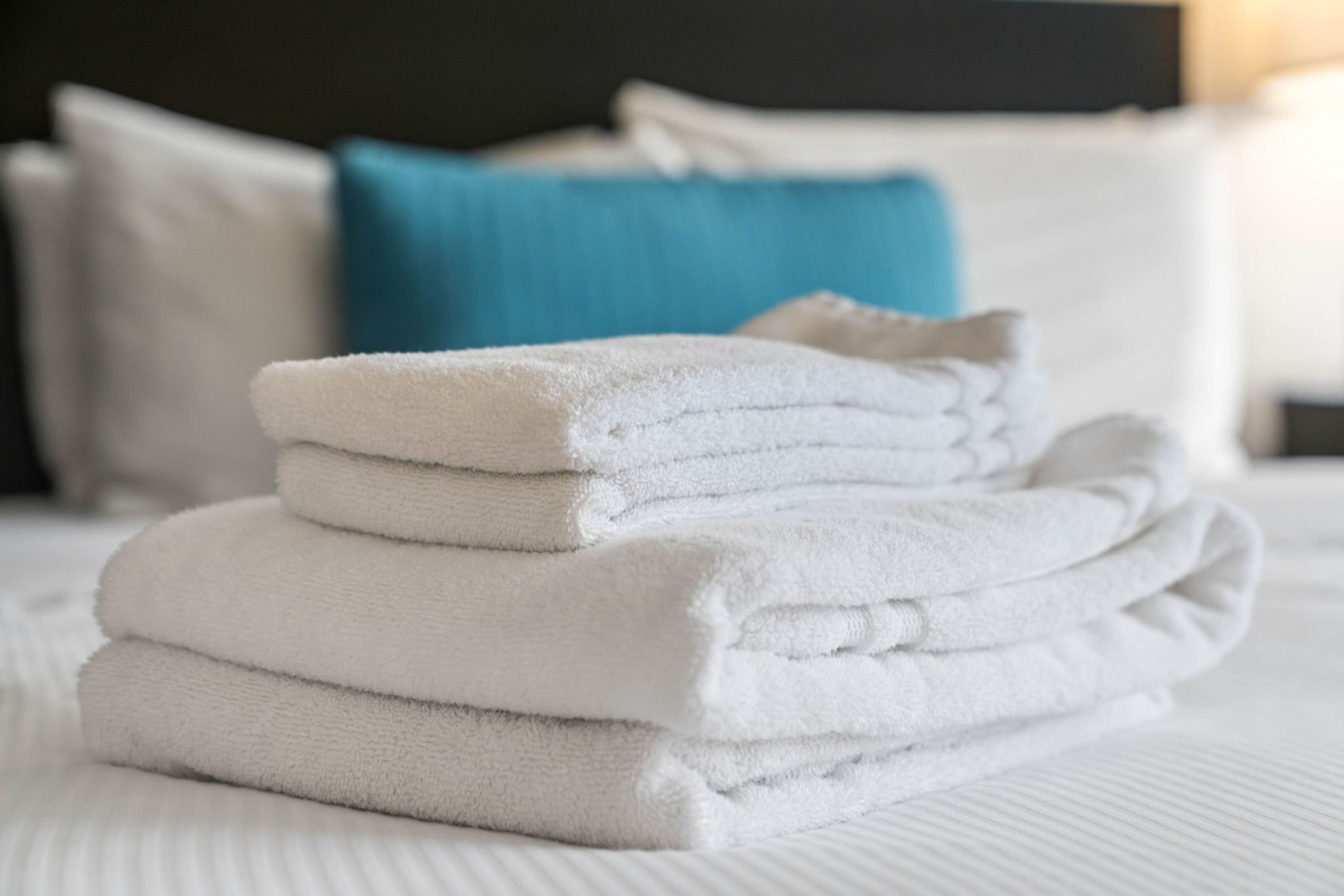 A neatly stacked pile of white towels on a bed with white bedding and a blue pillow in the background.