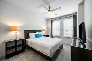 A modern hotel room with a bed, nightstands, a TV, a ceiling fan, and a window with blinds. - (S1BC) Great Location, Peaceful 1-BR Near Colony Square (Atlanta)
