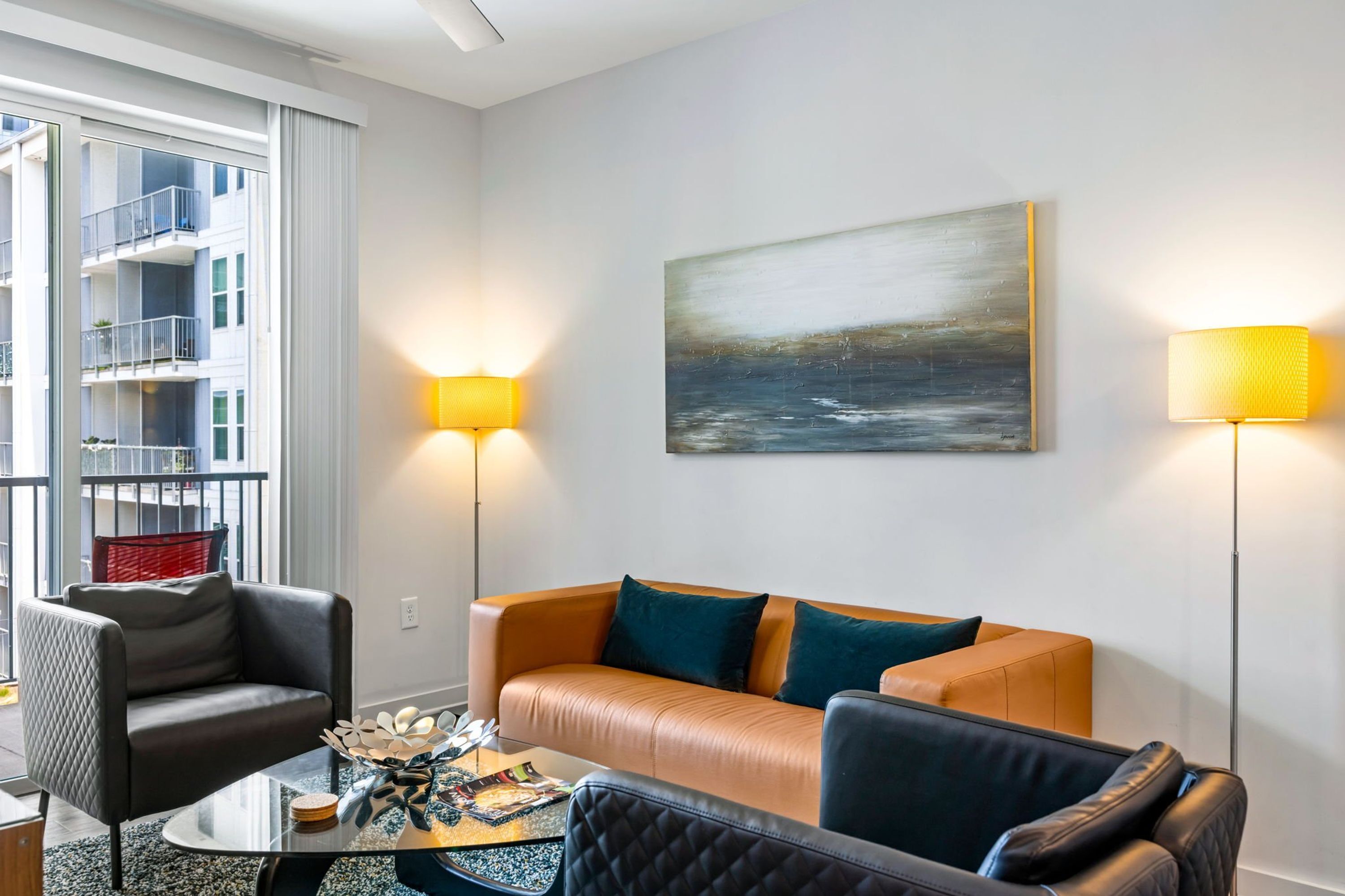A modern living room with a leather sofa, a glass coffee table, and a black armchair. There are two floor lamps and a painting on the wall.