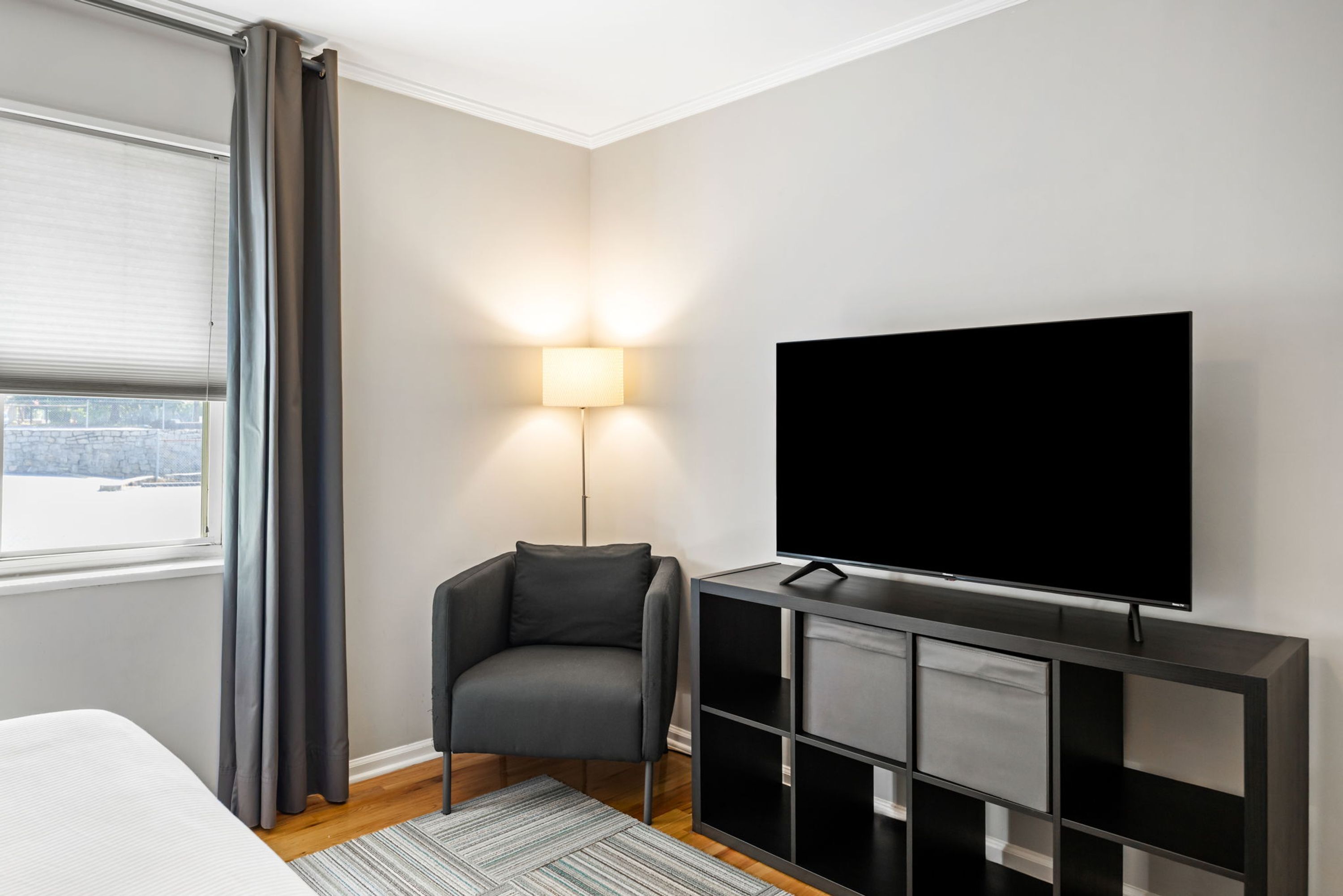 Une chambre d’hôtel moderne dotée d’une grande télévision à écran plat, d’un fauteuil gris, d’une table de chevet et d’une fenêtre avec des rideaux.