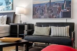 Ett modernt hotellrum med en svart lädersoffa, en vit säng, utsikt över staden och en lampa på ett sidobord.