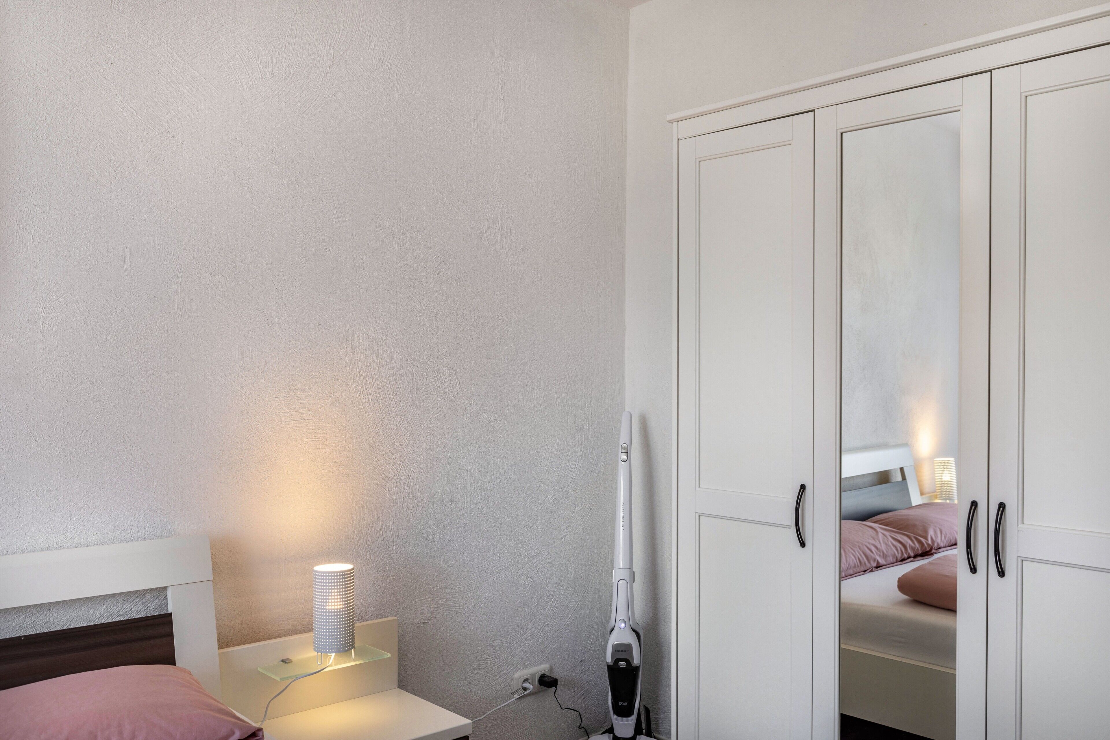 A bedroom with a bed, bedside lamp, nightstand, and a vacuum cleaner.