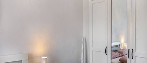 A bedroom with a bed, bedside lamp, nightstand, and a vacuum cleaner.