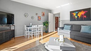 Ein modernes Wohnzimmer mit einem grauen Sofa, einem Couchtisch und einem Fernseher. Es gibt einen Essbereich mit Tisch und Stühlen sowie eine Pflanze auf dem Boden.