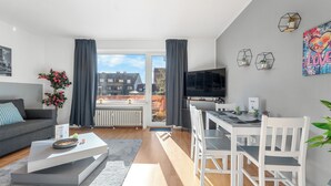 Ein modernes Wohnzimmer mit einem grauen Sofa, einem weißen Couchtisch und einem Essbereich mit weißem Tisch und Stühlen. Ein Fernseher ist an der Wand montiert, ein Fenster mit Vorhängen und Blick auf Gebäude im Freien.