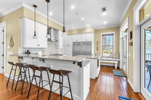 A kitchen with white cabinets, a granite countertop, and a stainless steel refrigerator. There are four stools at the island and a bench in the dining area. - Sailor's Cove by Lake Martin Vibe, 3 bedrooms, 2.5 baths, pets ok! (Dadeville)