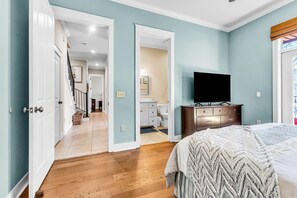 A bedroom with a bed, a TV, and a dresser. There is a hallway leading to a bathroom and a staircase.