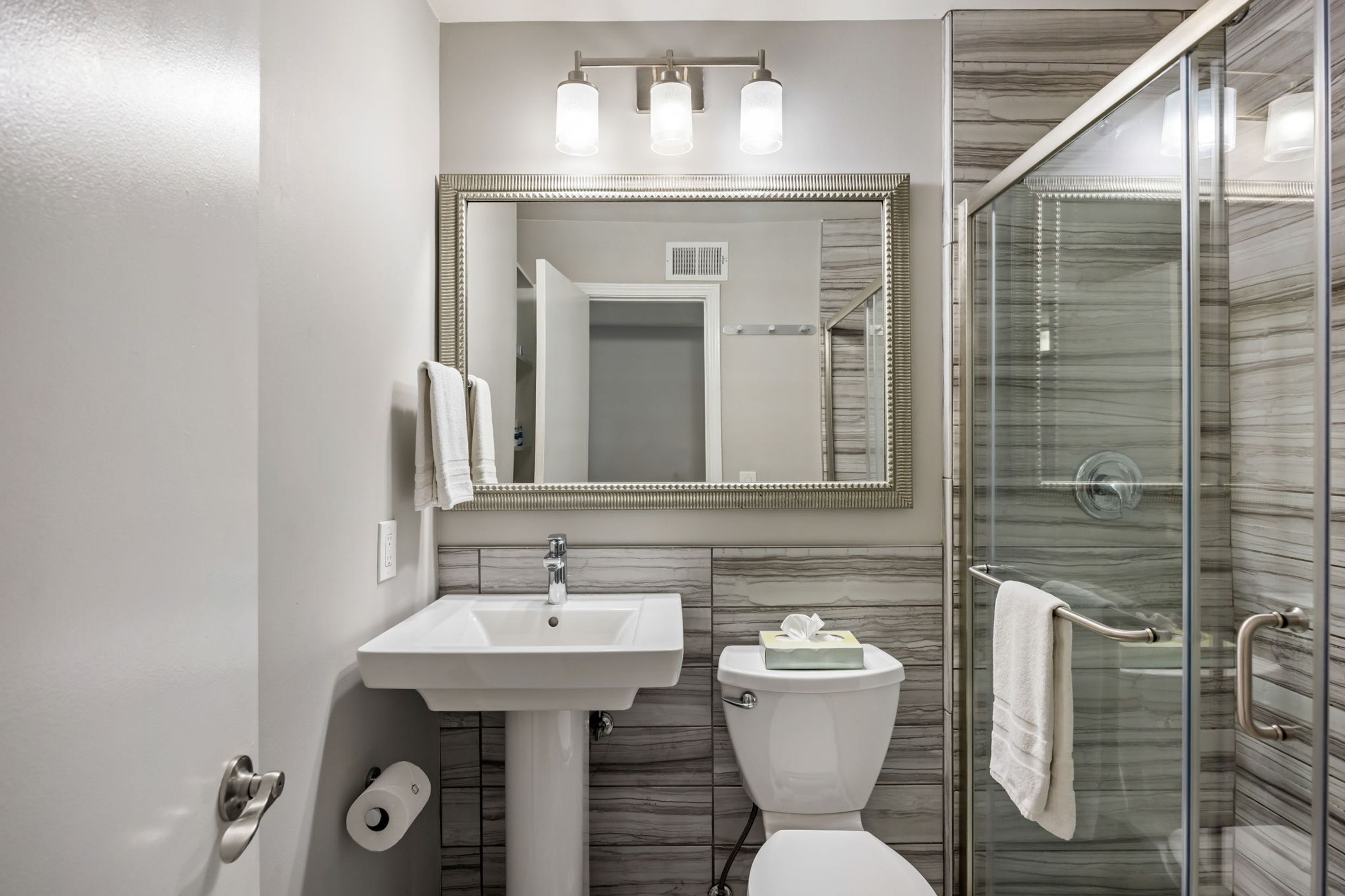 A bathroom with a white sink, a toilet, a glass shower enclosure, and a mirror with lights.