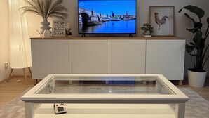 Een moderne woonkamer met een flatscreen televisie bevestigd aan een witte kast, een salontafel met glazen blad, een bank en een grote potplant.
