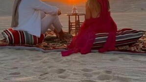 Twee personen zitten op een deken naar de ondergaande zon op het strand gericht.